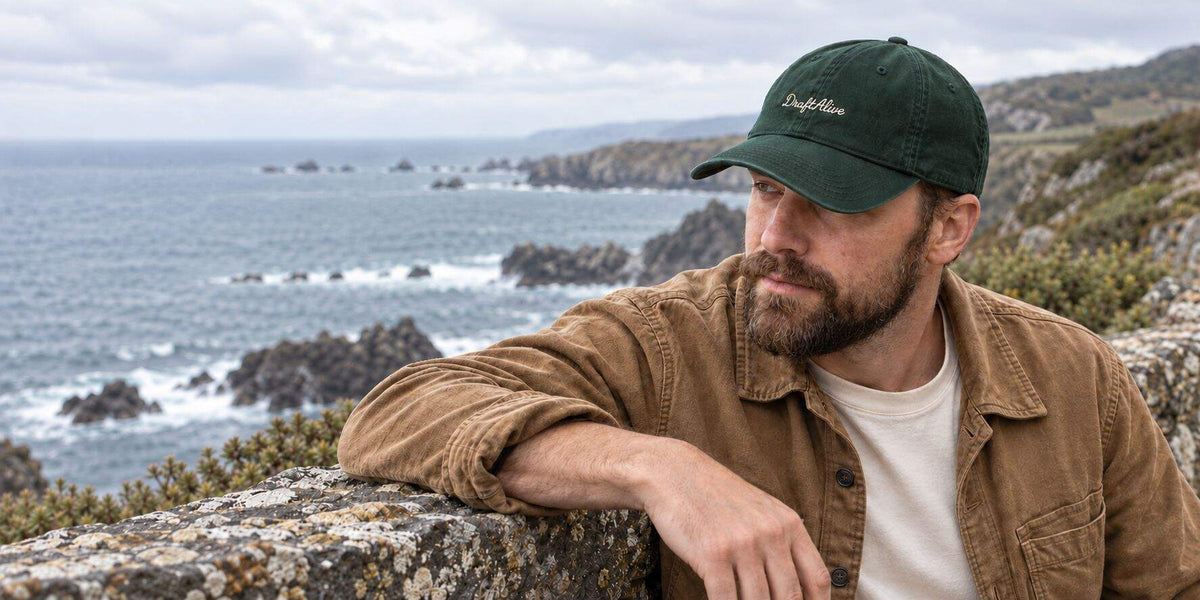 A man in an embroidered forest green cap resting on a stone wall above the sea